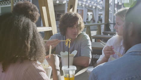 young man talking to his friends, while sitting around an outdoor table and eating street food