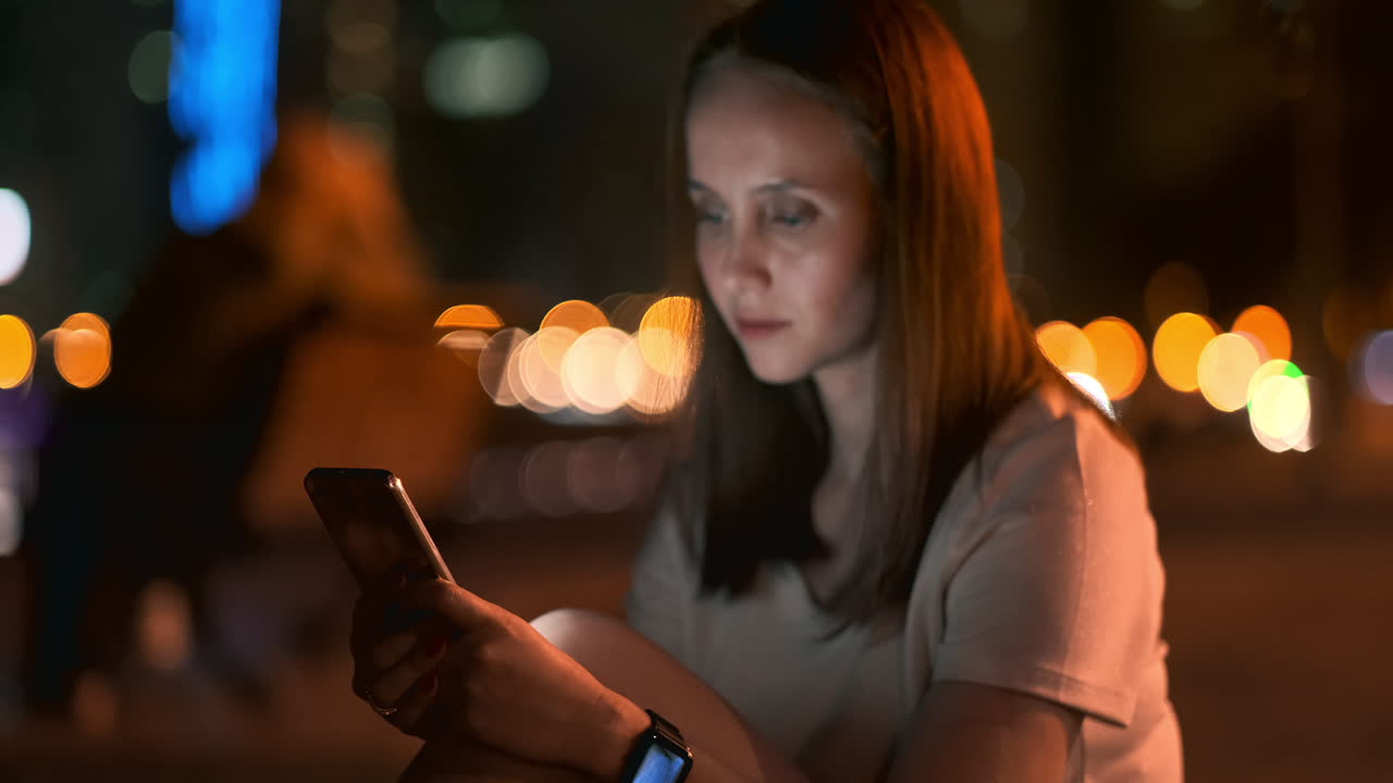 A young woman looks into the smartphone and writes text messages on the Internet against the backdrop of the night city. Girl businessman working on vacation remote work via mobile phone. Gadget addiction