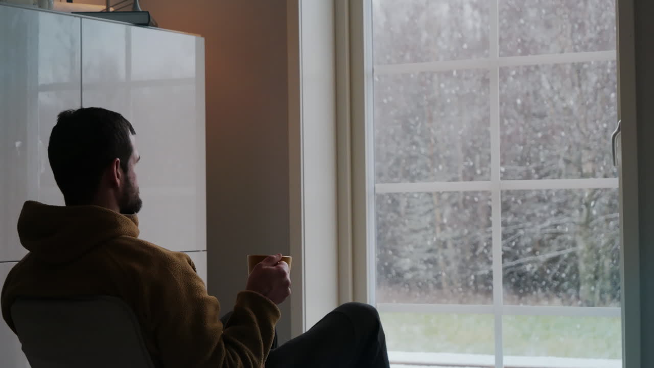 uomo che beve caffè mentre guarda la neve cadere dalla finestra