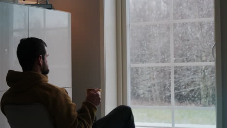 man-drinking-coffee-while-watching-the-snow-fall-through-the-window