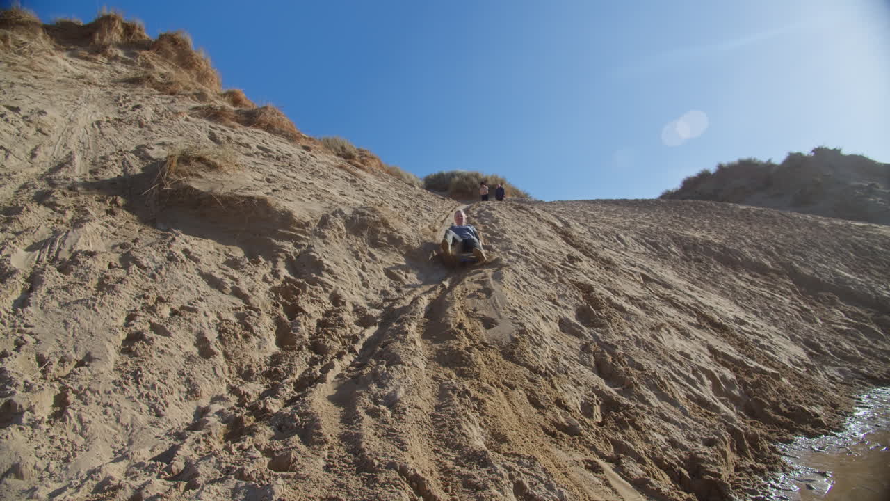 Woman sledging down sand dune having fun at Crantock Beach, wide shot