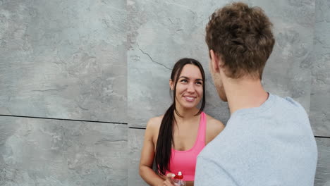 hermosa corredora sonriente apoyada contra una pared y hablando con su novio parado frente a ella mientras se toman un descanso durante una sesión de entrenamiento en la ciudad