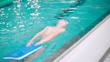 boy is doing backstrokes in indoor swimming pool while holding a paddle board