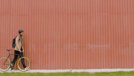 Young-American-Man-In-Formal-Clothes-Walking-With-His-Bike,-Then-Stopping-And-Looking-At-The-Camera-In-Front-Of-A-Prefab-Metal-Building