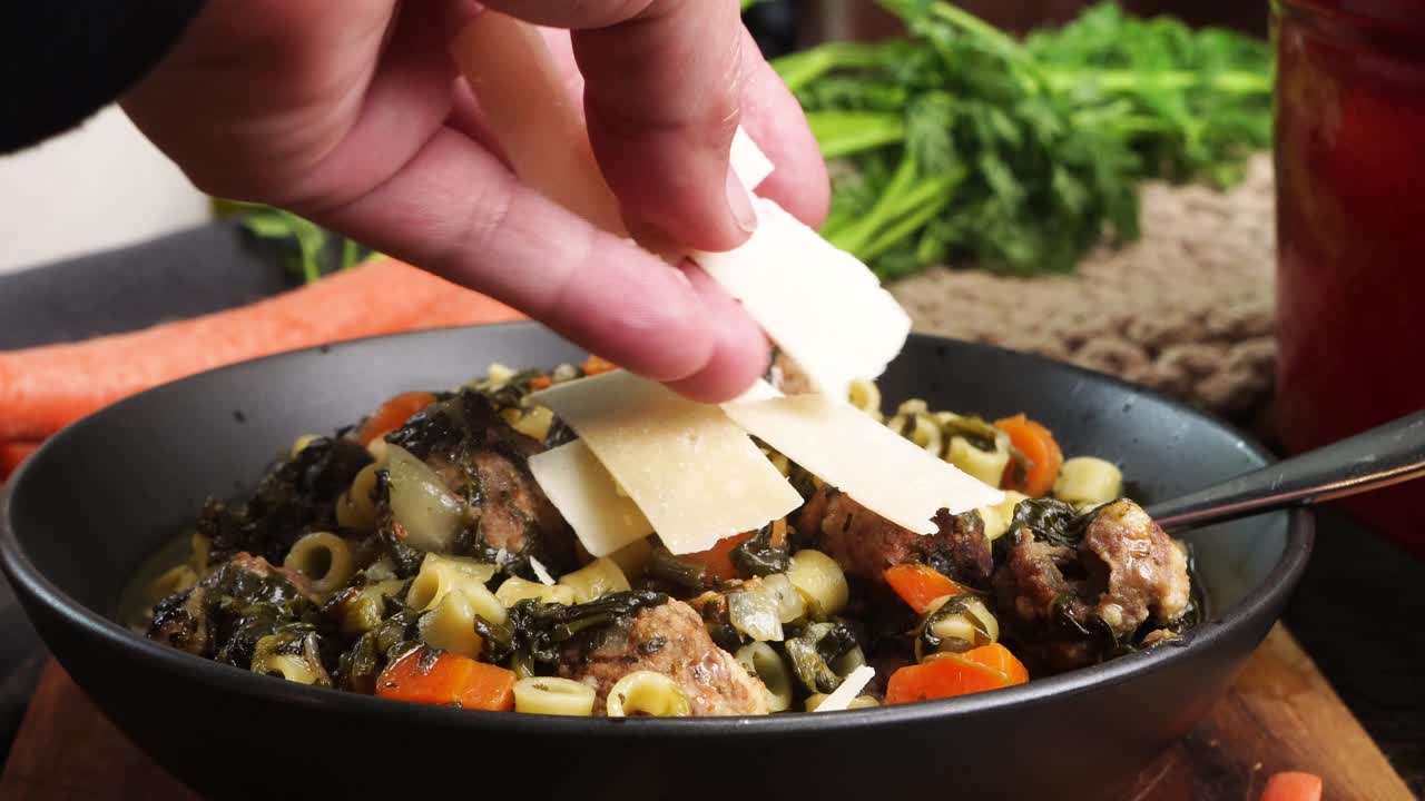 Cooking Italian Wedding Soup in the kitchen for dinner. Adding shredded parmesan cheese to the top of the soup as a garnish. Soup in a black bowl on a wood cutting board surrounded by fresh carrots.