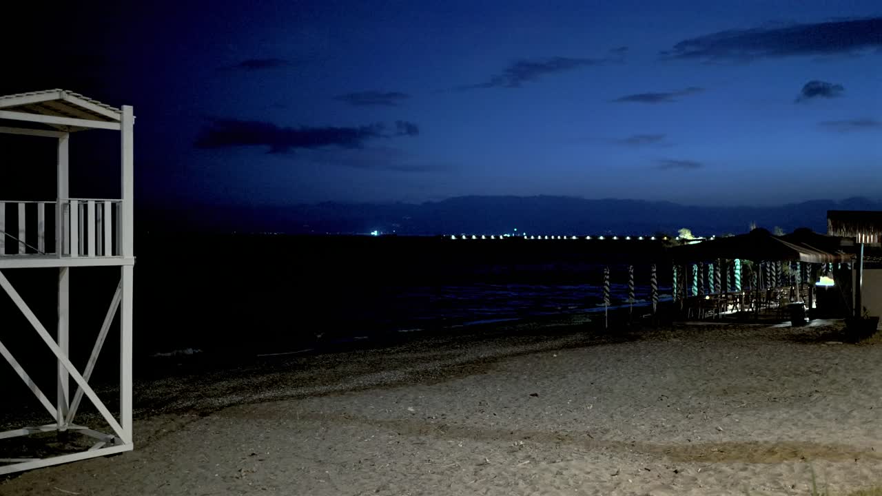 Wide panoramic pan view of empty beach at seashore of Kalamata during dusk, life guard tower and boat in frame, pier and city lights on background 4K