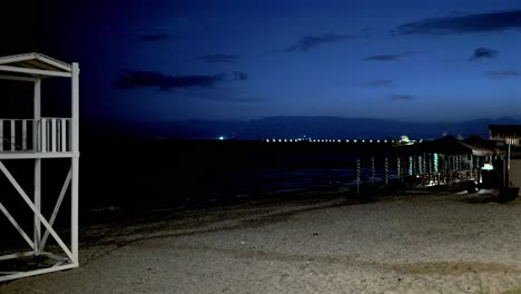 wide panoramic pan view of empty beach at seashore of kalamata during dusk, life guard tower and boat in frame, pier and city lights on background 4k