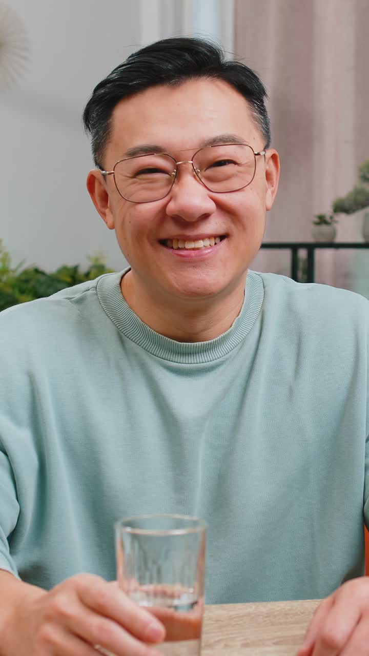 목마른 아시아 남자가 집 테이블에 앉아 천연 물 한 잔을 들고 물을 마시며 필터링 된 물을 마신다.