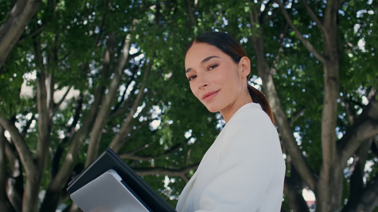 성공한 사업가 여성이 녹색 나무 근처에 서서 자신감 있게 카메라를 보고 있습니다.