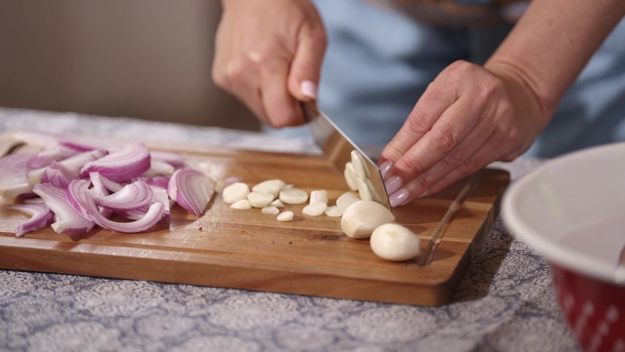 Close-up tangan mengiris siung bawang putih segar dan bawang merah di atas talenan kayu. Persiapan masakan rumahan dengan bahan-bahan segar untuk hidangan rumahan sederhana