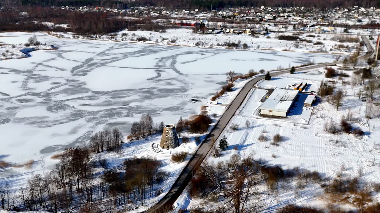 A frozen lake marked with natural ice patterns sits beside a rural tower and buildings on the edge of town, surrounded by snowy fields and bare trees in a calm winter setting from above.