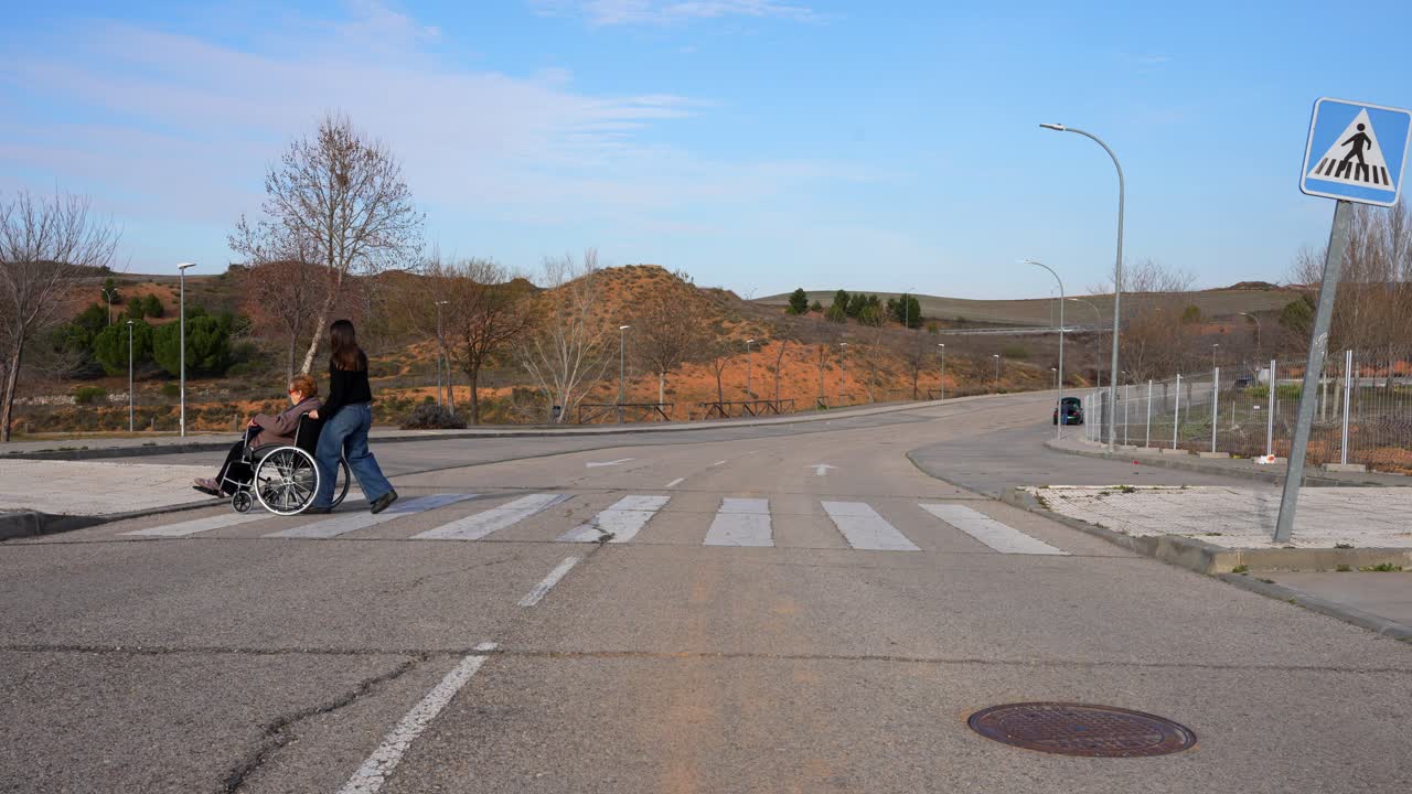 Young woman carrying elderly woman in wheelchair across crosswalk