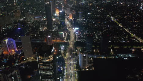 Wide-aerial-view-of-multi-lane-highway-traffic-at-night-in-modern-Jakarta-city-center-with-tall-skyscrapers