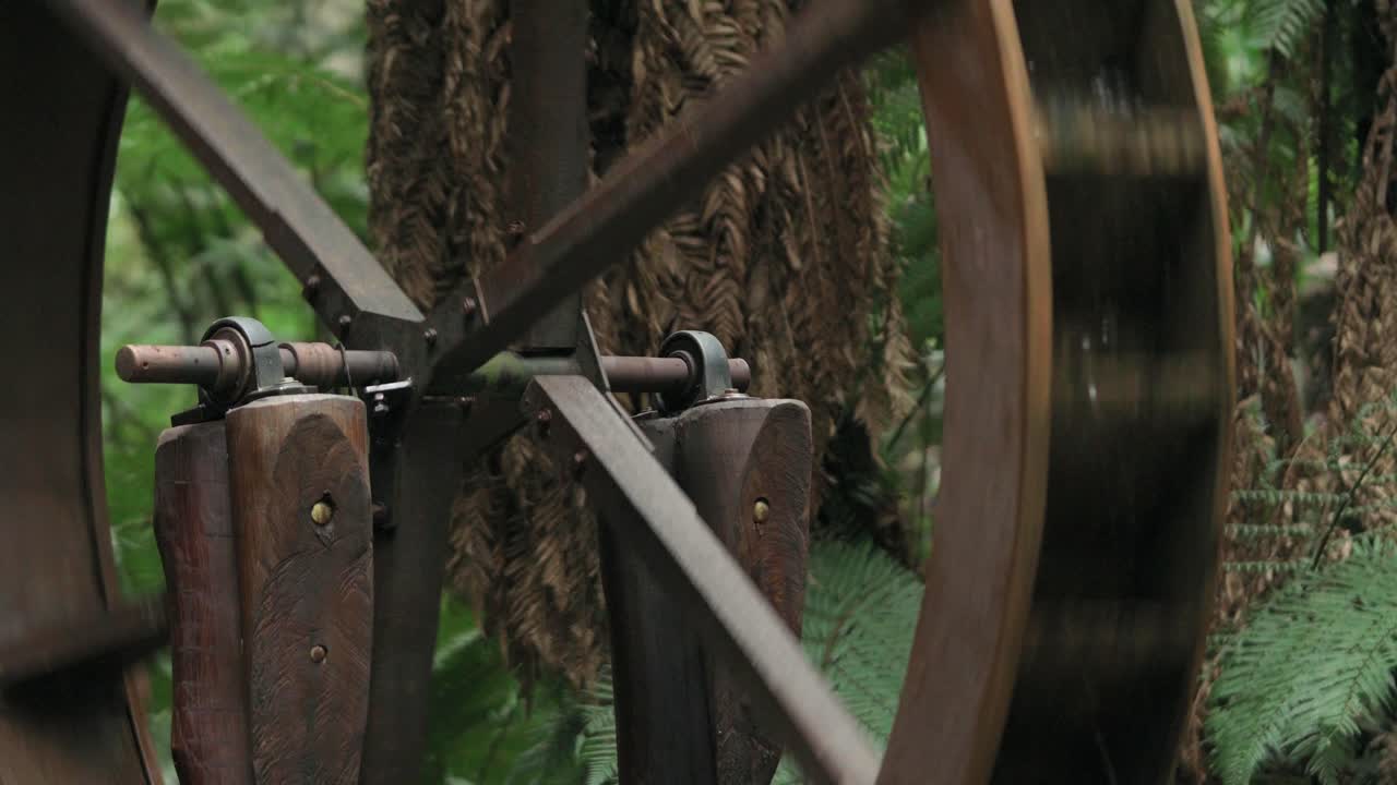 Tangkapan close-up detail yang berfokus pada jari-jari kayu dan struktur pedesaan dari kincir air tradisional di lingkungan hutan hijau