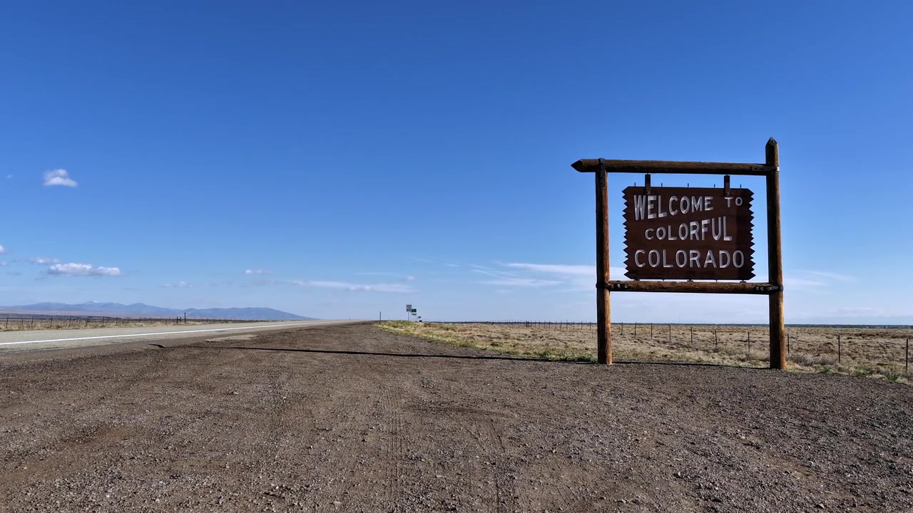 Timelapse atmosférico en 4K del icónico y rústico letrero de carretera "Welcome to Colorful Colorado" a lo largo de una carretera desolada. Vastas llanuras extensas y un cielo azul vibrante crean una estética de viaje cinematográfica.