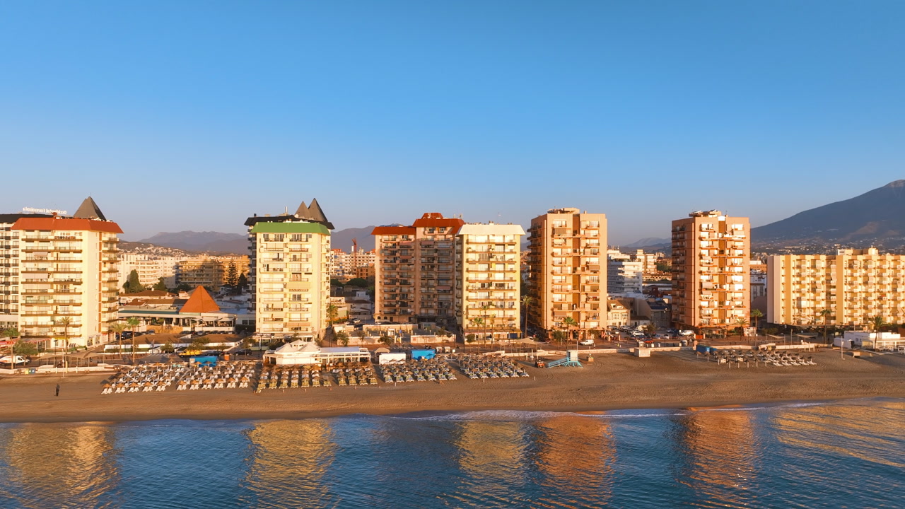 Flight reverses back revealing ten storey hotels and apartment buildings, waterfront promenade and quiet golden brown beach at day break. Early morning sun casts golden glow over the scene Fuengirola
