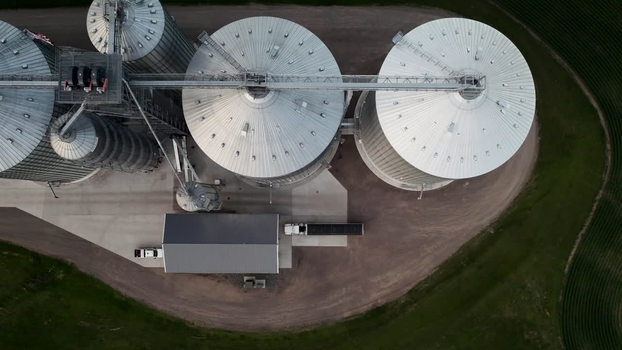 Overhead drone footage of large circular grain silos at an agricultural storage facility with a grain truck driving alongside