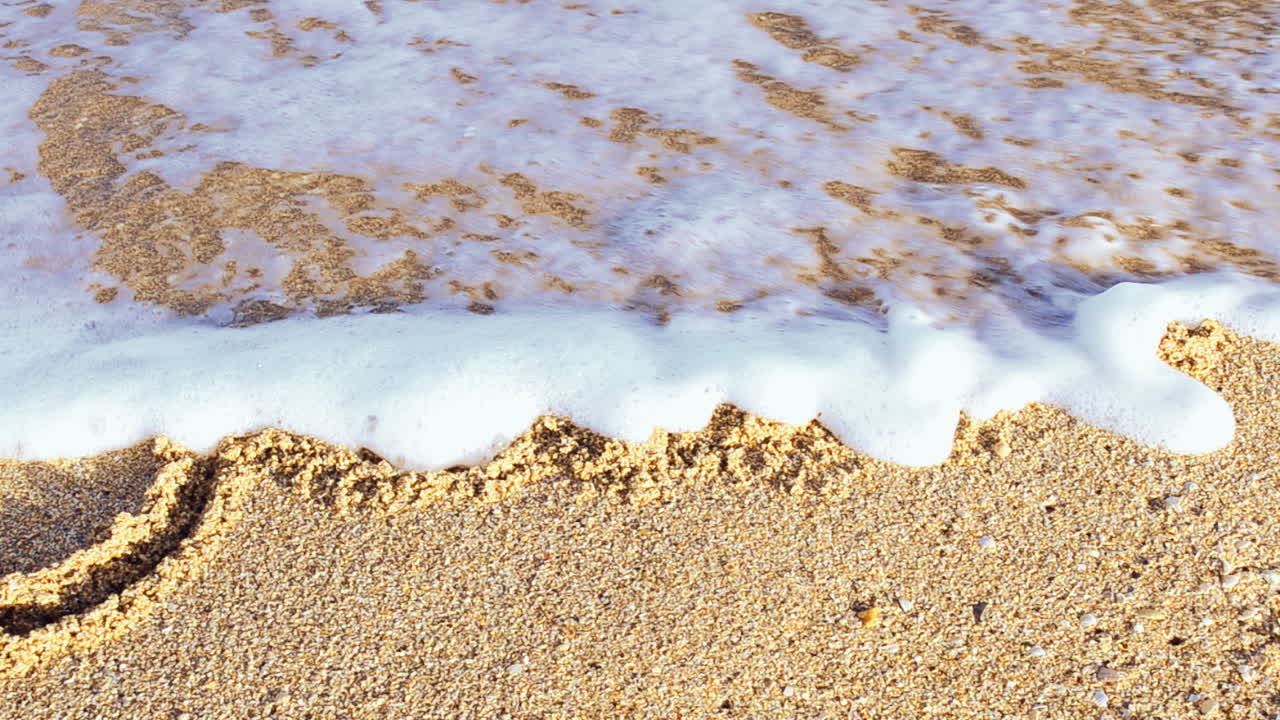 Iscrizione Giugno sulla sabbia. L'iscrizione viene cancellata da un'onda del mare. Riprese sulla spiaggia.