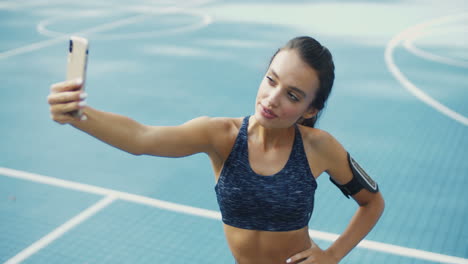 mujer fitness soplando un beso de aire y tomándose selfie en un smartphone en una cancha deportiva en un día de verano
