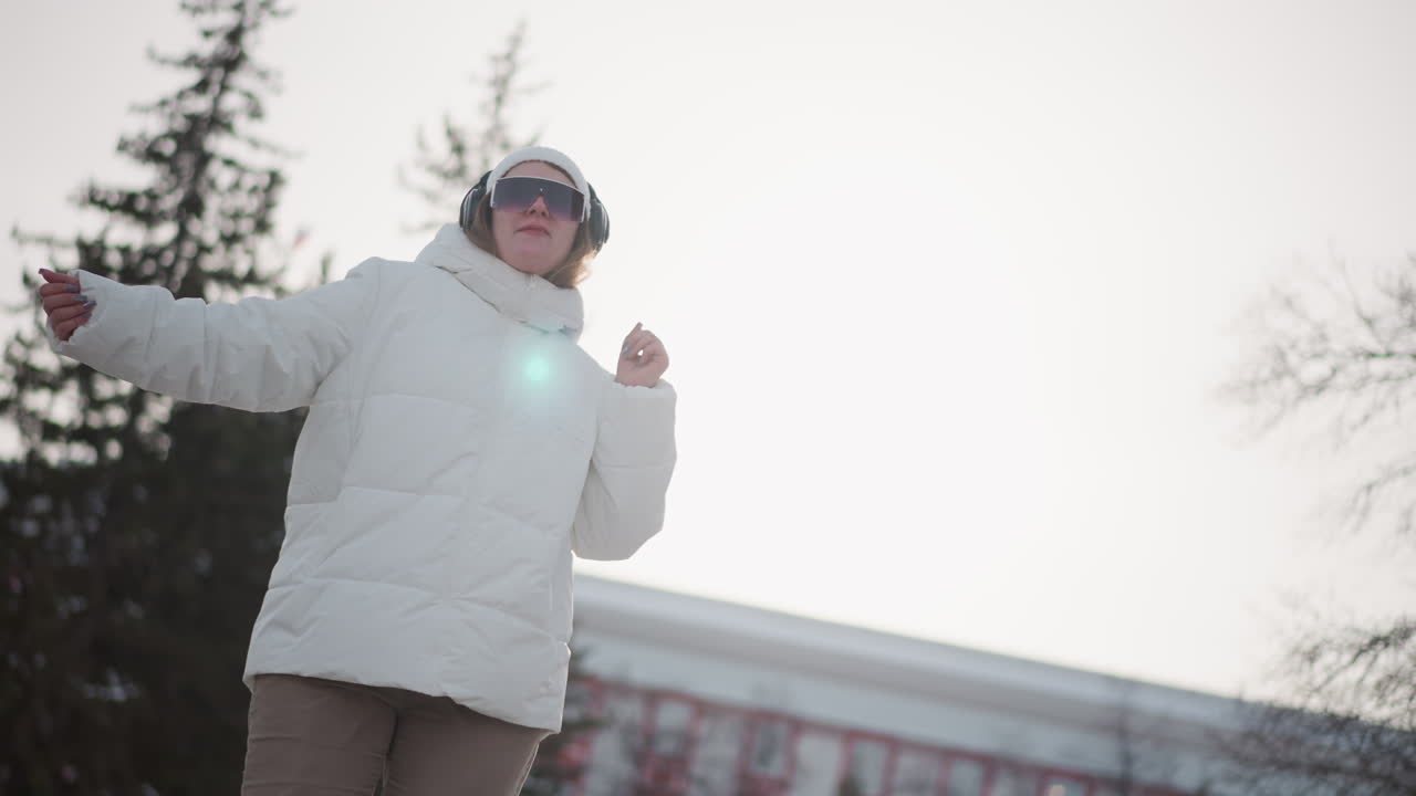 Woman wearing white puffer jacket, beanie, black goggles and headphones dancing energetically and smiling while grooving with fluid hand gestures on snow dusted urban plaza to upbeat winter music