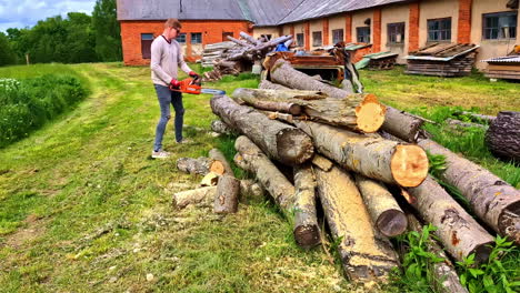 man cutting logs with chainsaw next to large woodpile in sunlit yard