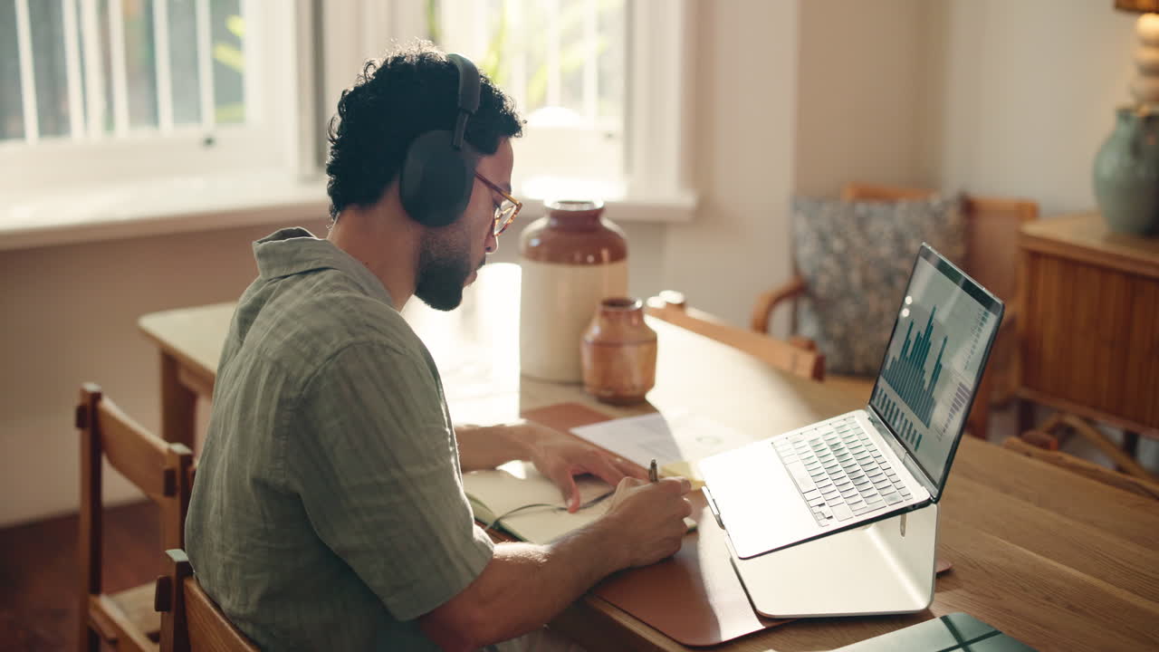 Uomo con le cuffie che lavora al laptop e prende appunti a casa
