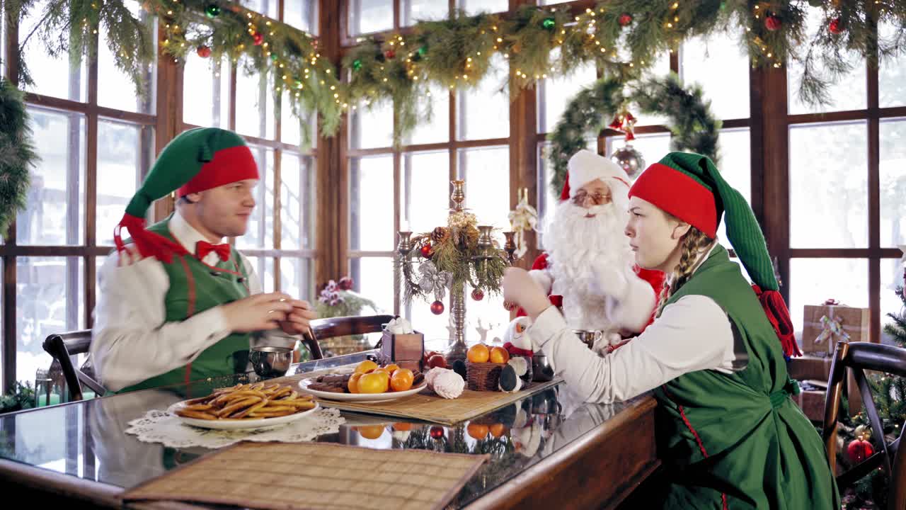 Elves and Santa Claus at the table at Christmas. Joyful fairy characters having great time together while eating sweets and cookies on New Year time.