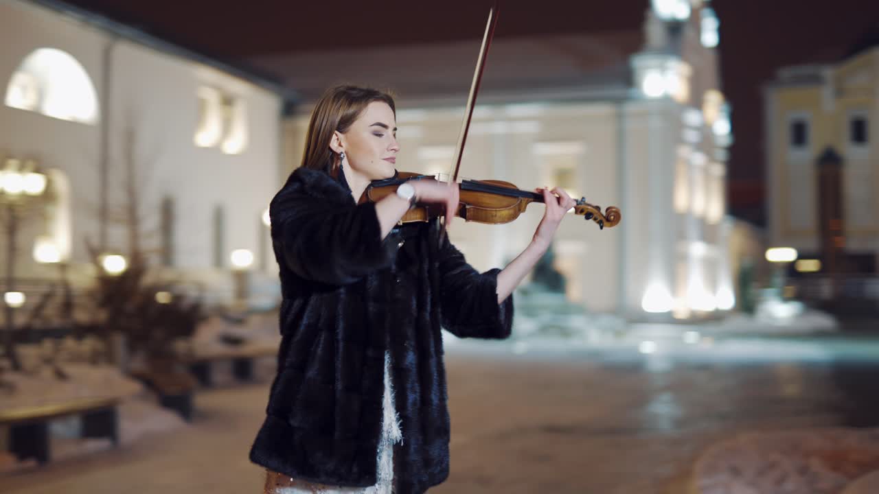 A talented girl performs composition on the instrument on the background of tall buildings in the city center. The musician plays the violin in the evening. Blurred background.