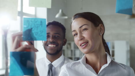 Businesswoman-Writing-Notes-On-The-Window-Pane-While-Talking-To-An-Colleague