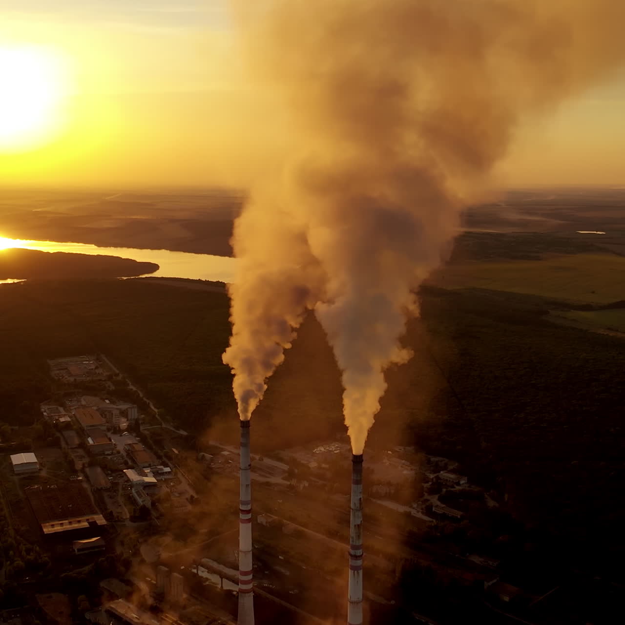 Two pipes with thick smoke. Industrial factory surrounded by nature in the evening. Air pollution. Aerial view. Slow motion camera.