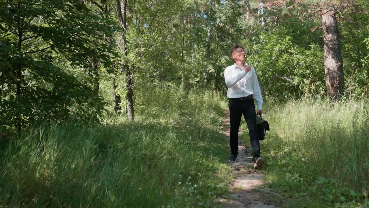 Tall man in white shirt and black trousers carrying backpack walking slowly on forest path, pointing hand while observing plants surrounded by lush greenery and sunlight streaming through trees