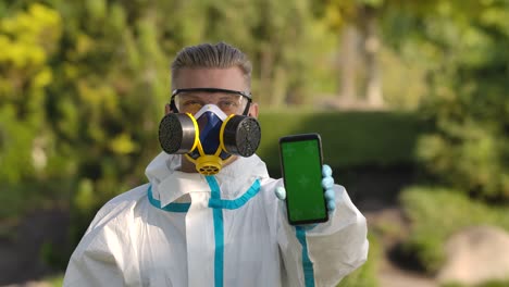 retrato hombre joven en un traje de protección, gafas, un respirador y guantes sostiene un teléfono inteligente con pantalla verde y tecla de croma con posición vertical de la pantalla y señala con un dedo.