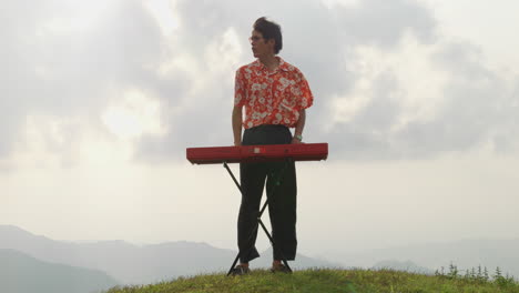 Asian-guy-playing-keyboard-at-mountain-in-Vietnam,-wide-angle-front-handheld-slow-motion