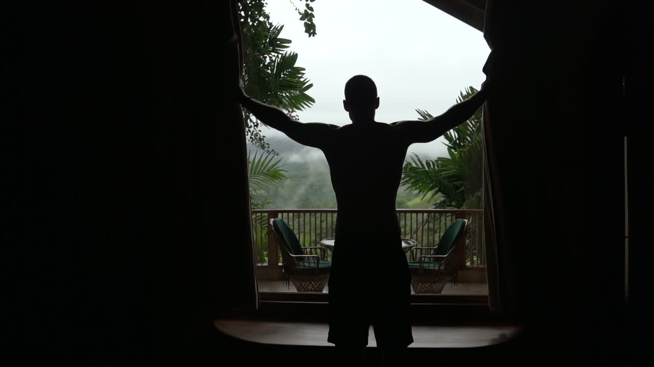 Silhouetted mann opens blinds to reveal balcony in foggy rain forest, slow motion