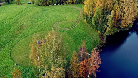 Toma-Aérea-De-Arriba-Hacia-Abajo-Del-Campo-De-Hierba-Con-Sauna-De-Barril-Junto-Al-Lago-Natural-Durante-El-Día---Paisaje-Idílico-En-La-Naturaleza