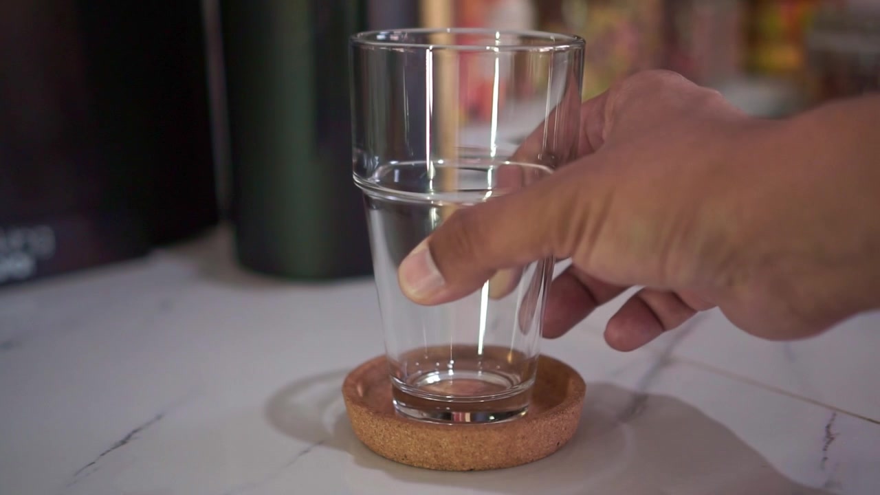 Mano che appoggia un bicchiere trasparente su un sottobicchiere di legno, primo piano. Concetto di preparazione domestica, stile di vita legato alle bevande, clip di filmato di repertorio.