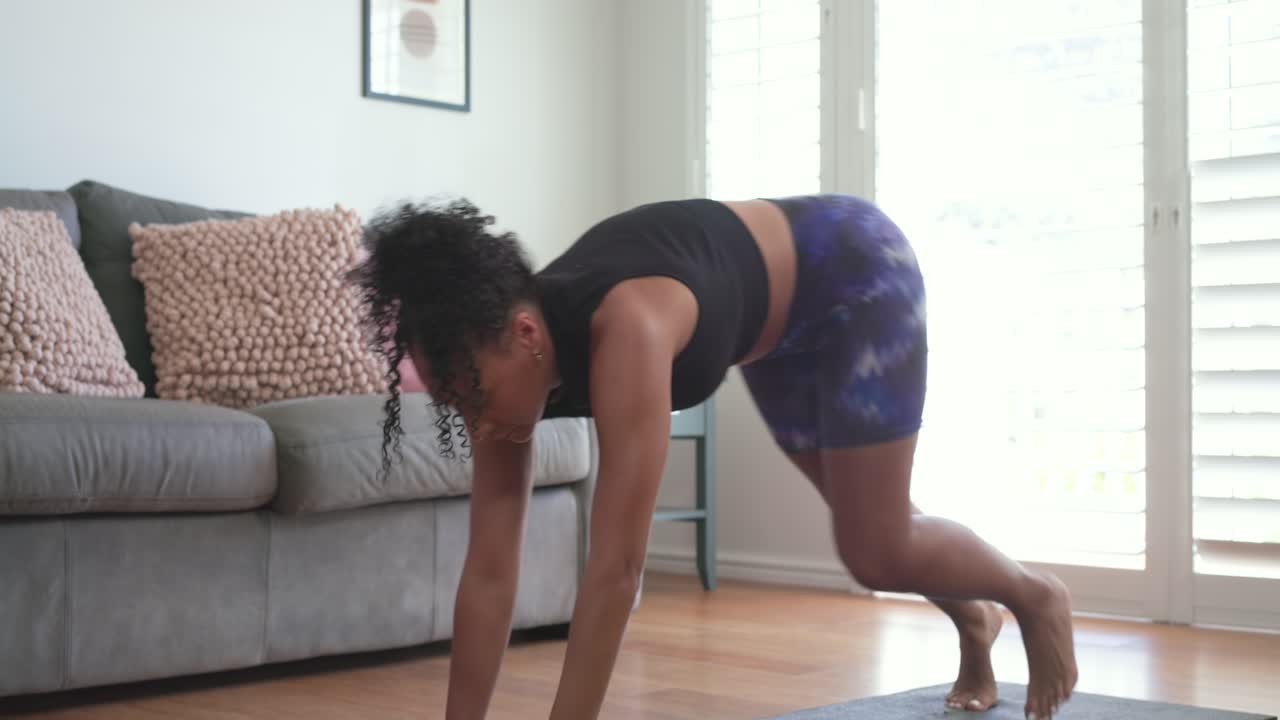 African woman starting low lunge, flowing into twist on home mat, in sportswear for workout