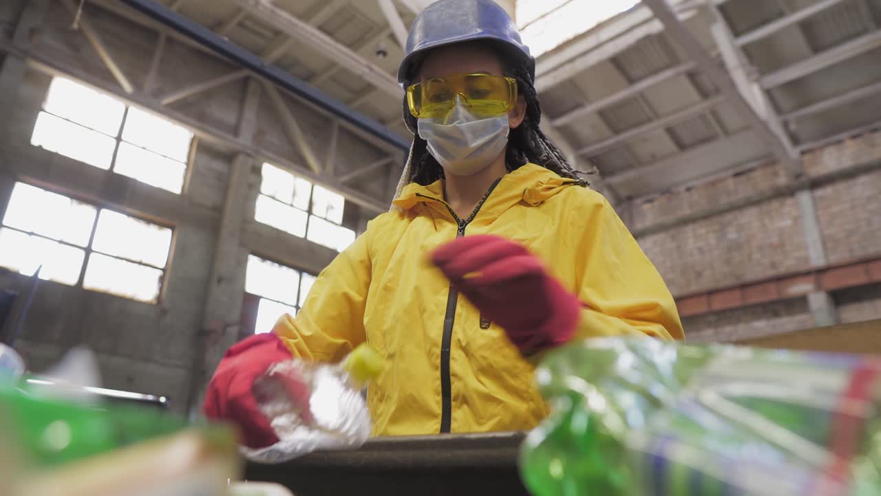 mujer voluntaria con chaqueta amarilla y gafas de protección transparentes, casco y máscara clasificando botellas de plástico usadas en el reciclaje