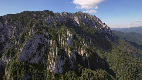 Las-Majestuosas-Montañas-De-Piatra-Craiului-Bajo-Un-Cielo-Azul-Claro,-Frondosos-Bosques-En-El-Fondo,-Vista-Aérea