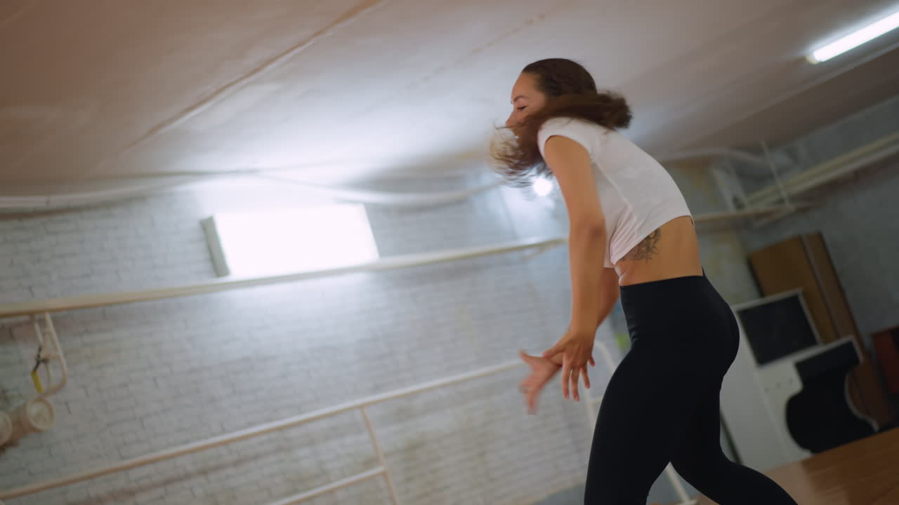 Vista posterior de uma mulher de blusa branca e leggings pretas dançando energicamente enquanto joga o cabelo dentro de um estúdio de fitness, mostrando ritmo, poder e graça durante um treino expressivo e confiante