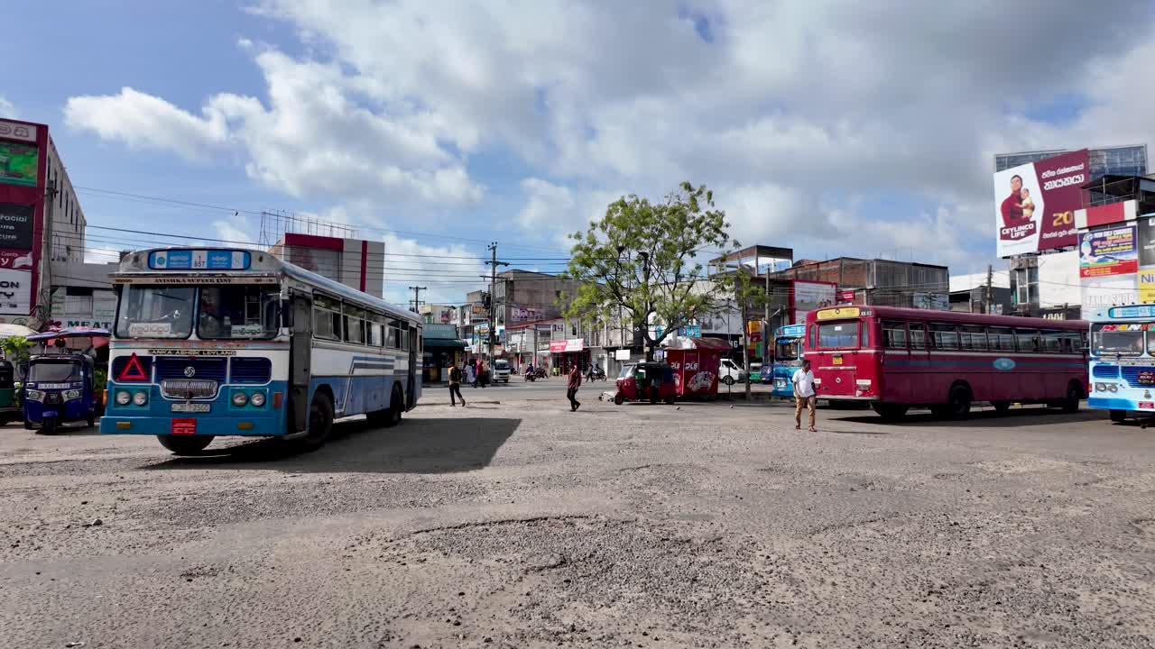 Local bus stop in Anuradhapura, Sri Lanka. The image captures buses, tuk-tuks, and people against a backdrop of urban buildings under a clear sky.