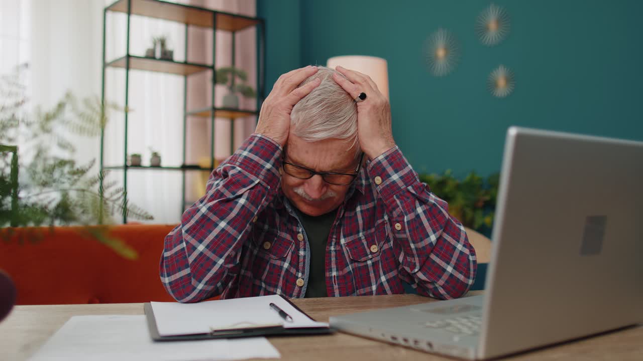 Senior man working on laptop computer making notes on sheet of paper upset by poor bad results