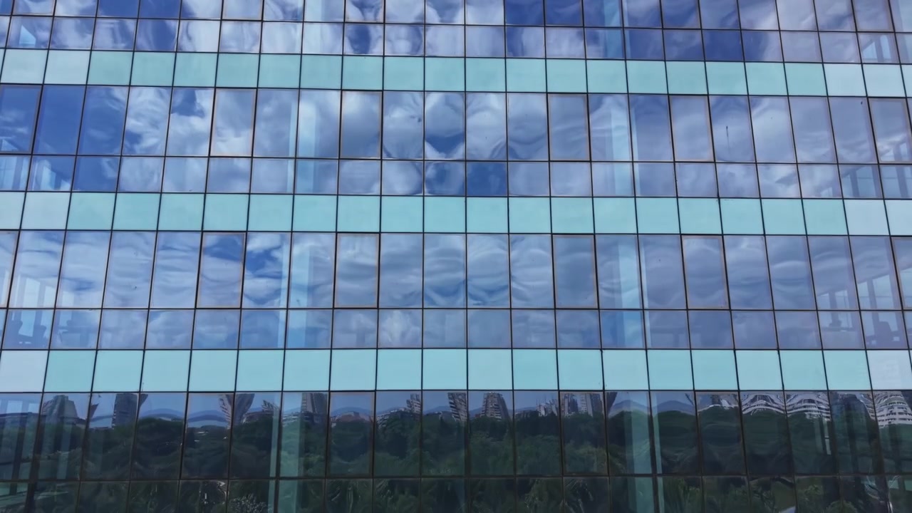 Edifício com vidro reflexivo mostrando nuvens e céu azul durante o dia em uma área urbana perto de vegetação.