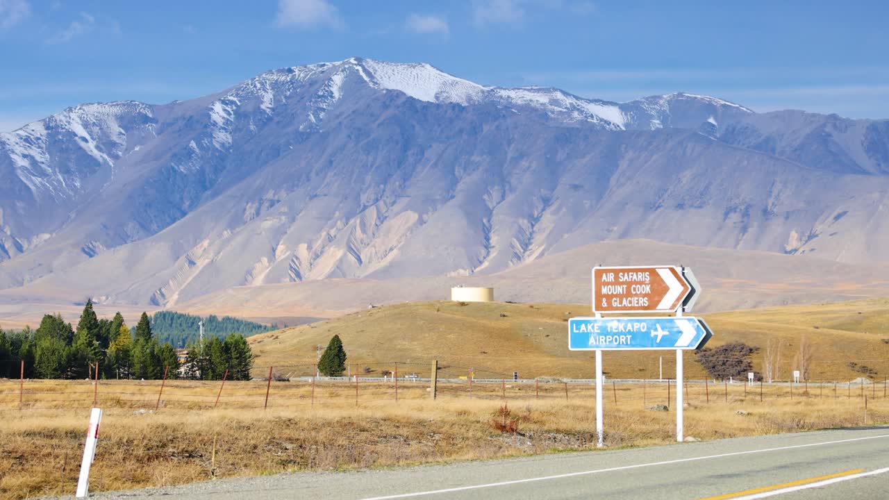 A silver car drives along a rural road past directional signs, with snow-capped mountains and dry grasslands under bright daylight and clear skies