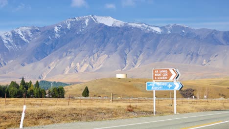 a silver car drives along a rural road past directional signs, with snow-capped mountains and dry grasslands under bright daylight and clear skies