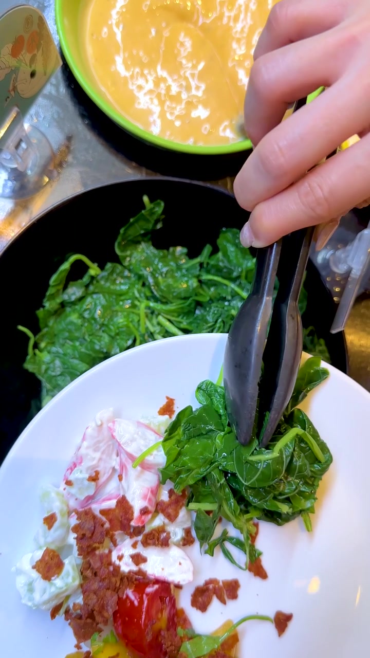 Una persona utiliza pinzas negras para colocar espinacas frescas en un plato en un bufé.