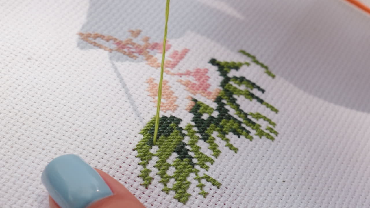 Close up designer embroidering fabric, needle passing through cross stitch pattern, blue nail visible, green and peach threads forming leafy design, precise work on white canvas under soft daylight