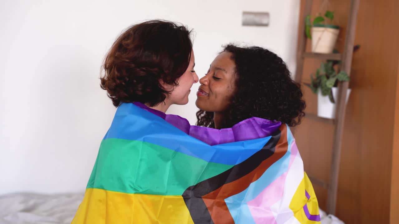 Happy diverse young friends with LGBT flag touching nose tips sitting in bed