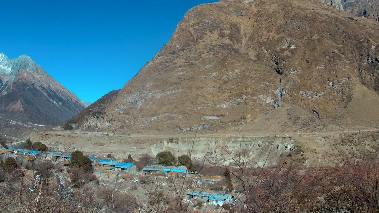 Daily life in the Himalayan region of Nepal. Locals carry wooden logs in a basket on their back to the village for firewood and livestock. Panning shot of Mount Manaslu and Samagaun village