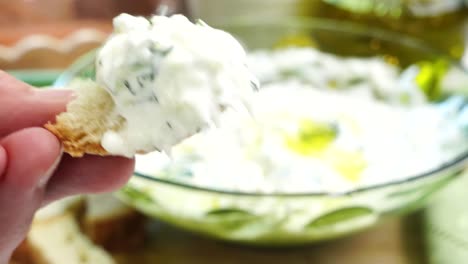 dipping bread into tzatziki sauce. making tzatziki greek or mediterranean sauce or dip for a party. dip or sauce in a green bowl on a wooden cutting board with dill, green napkin and a grater.